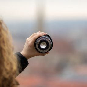 Curly-haired person holding a camera lens with a city view, offering a creative perspective.